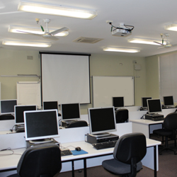 There are three rows of desks set up facing the front of the room. Two computers are at each desk. The front of the room has a whiteboard and a projector set up