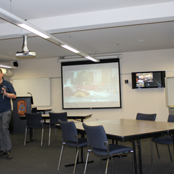 There are desks set up in squares around the room. The front of the room has a whiteboard and a projector set up.