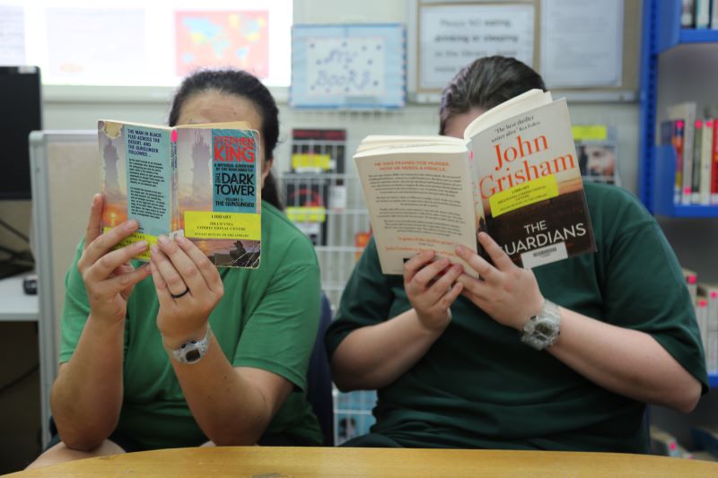 Two women dressed in green, seated at a table, and holding books in front of their faces