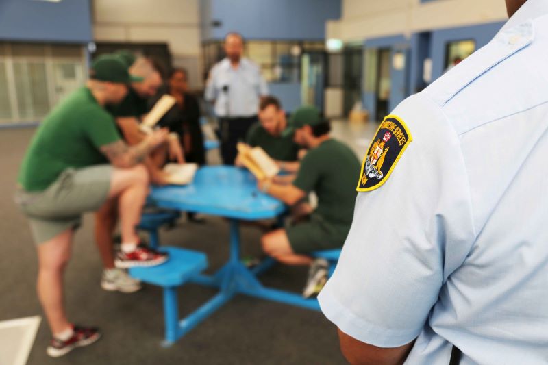 Corrective officer in foreground, blurred inmates gathered around a bench with books in background.