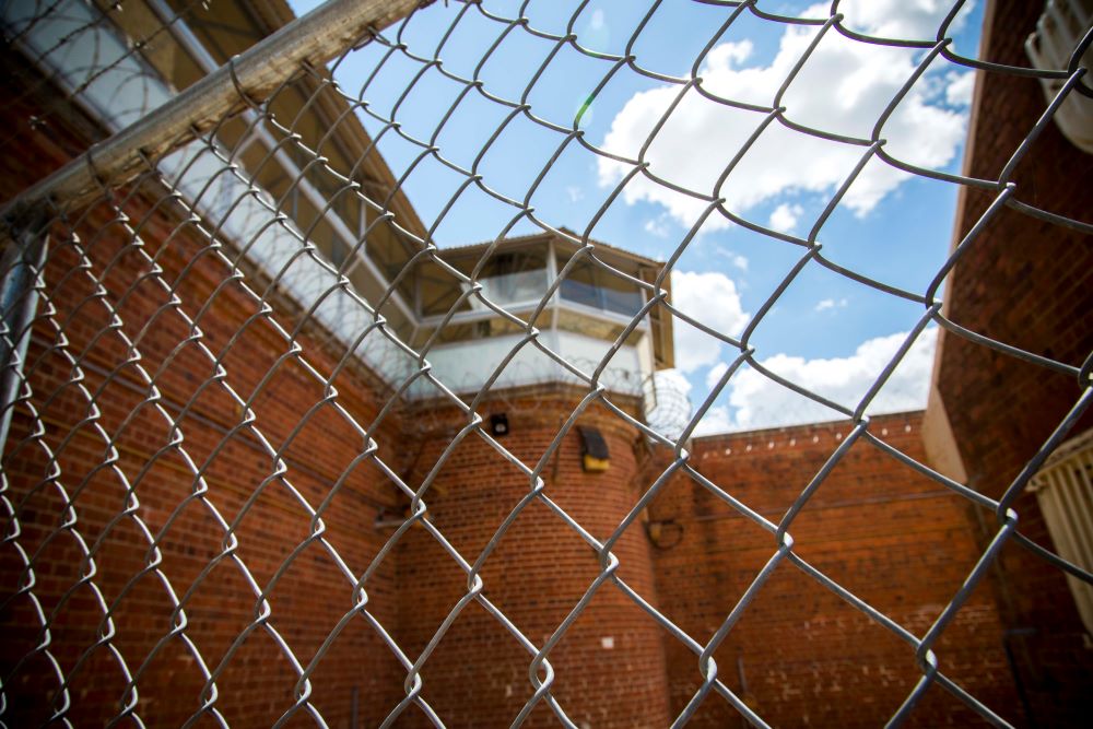 Mesh fence in foreground, prison tower and barbed wire in background