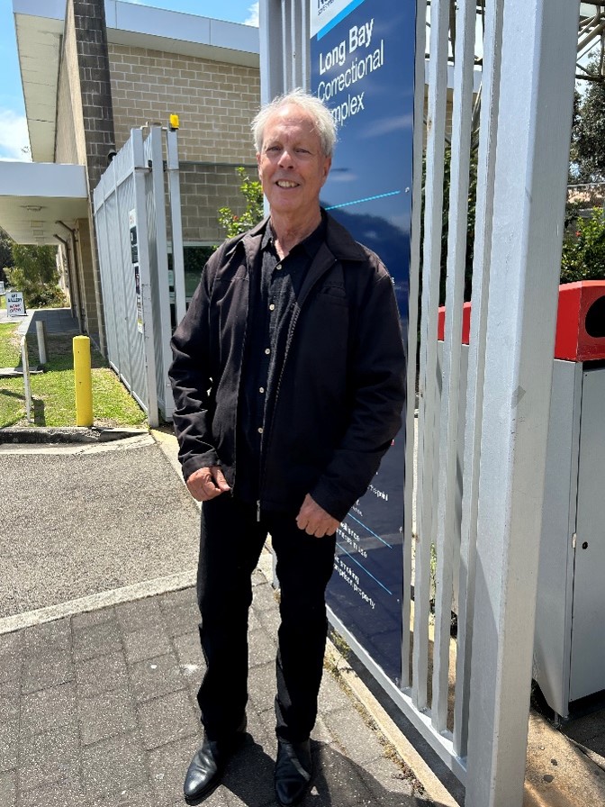 Murray Cook dressed in black jacket, pants and shoes standing in front of Long Bay gates. 