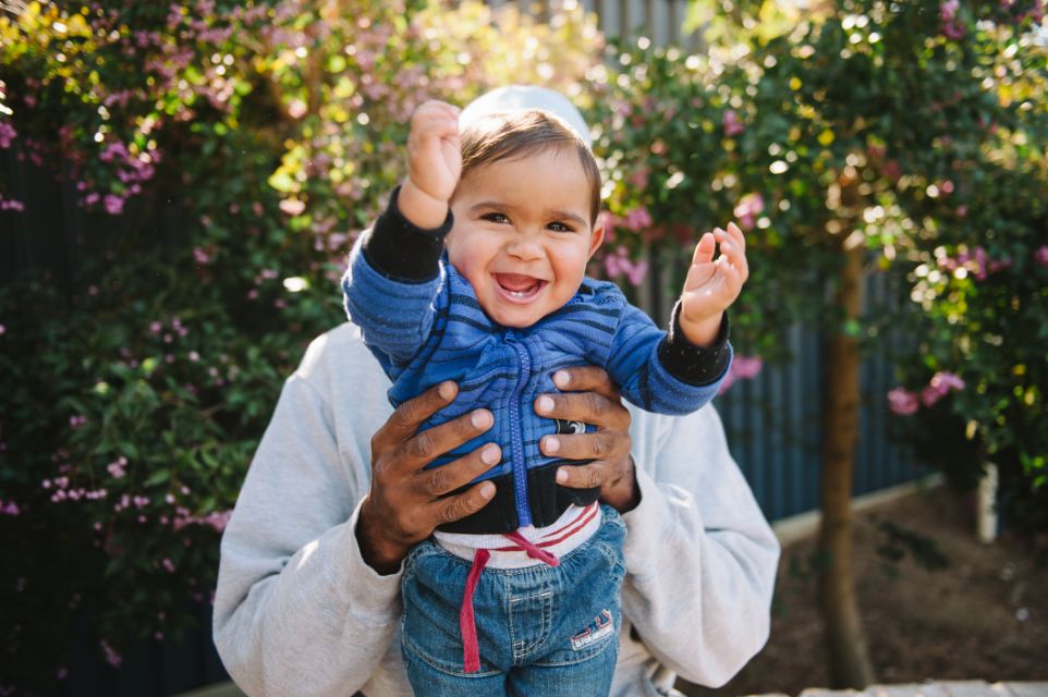 A person holding up a young boy who is making a funny face and smiling with his arms up
