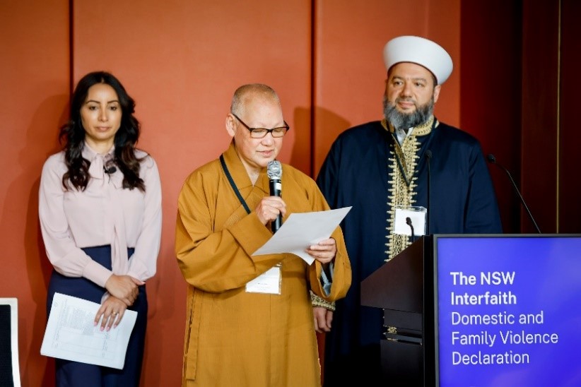 Three people standing on the stage at the front of the room. The one in the middle is reading a document while the other two are listening.