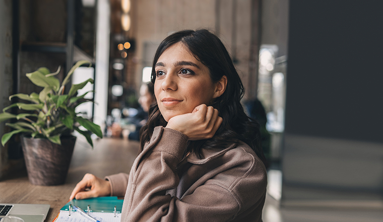 Young woman sitting at a desk in a public space, looking thoughtfully out the window, with a plant and laptop nearby.